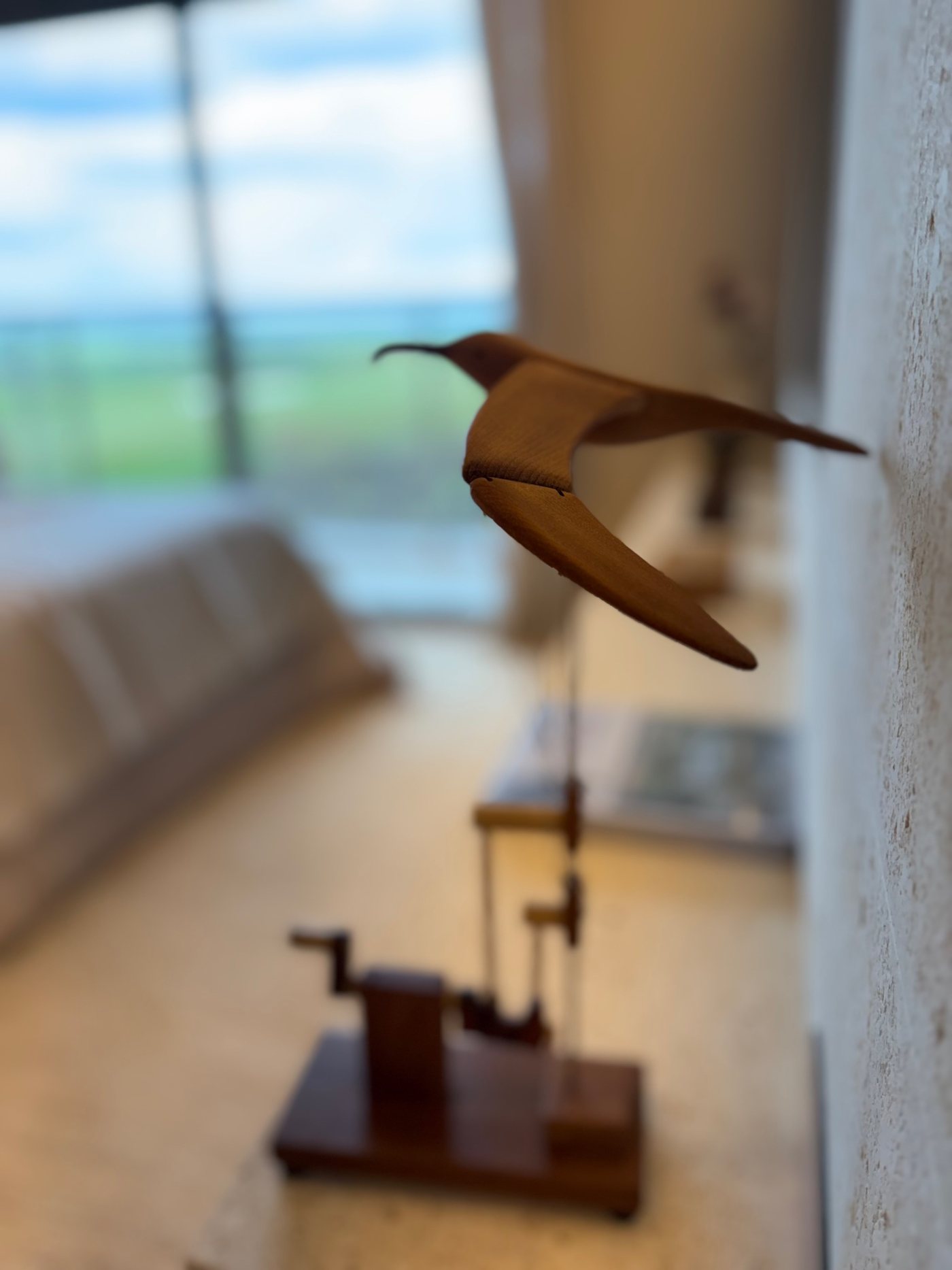 Pássaro de madeira em voo preso à parede, com a sala e a paisagem desfocadas ao fundo