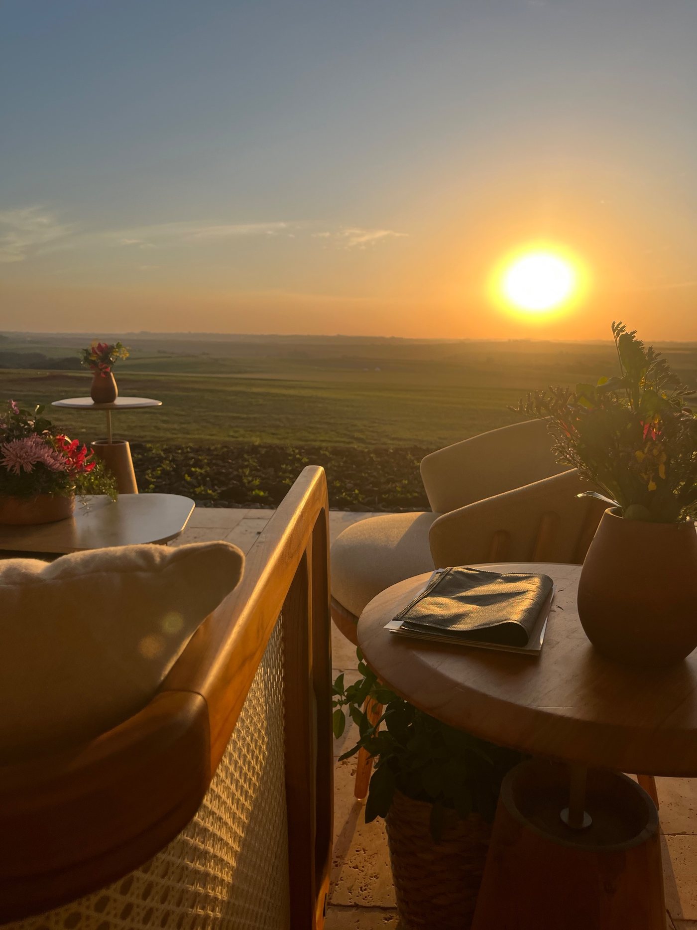 Varanda ao pôr do sol, com poltronas de madeira e palhinha, mesa com flores e a paisagem do campo ao fundo