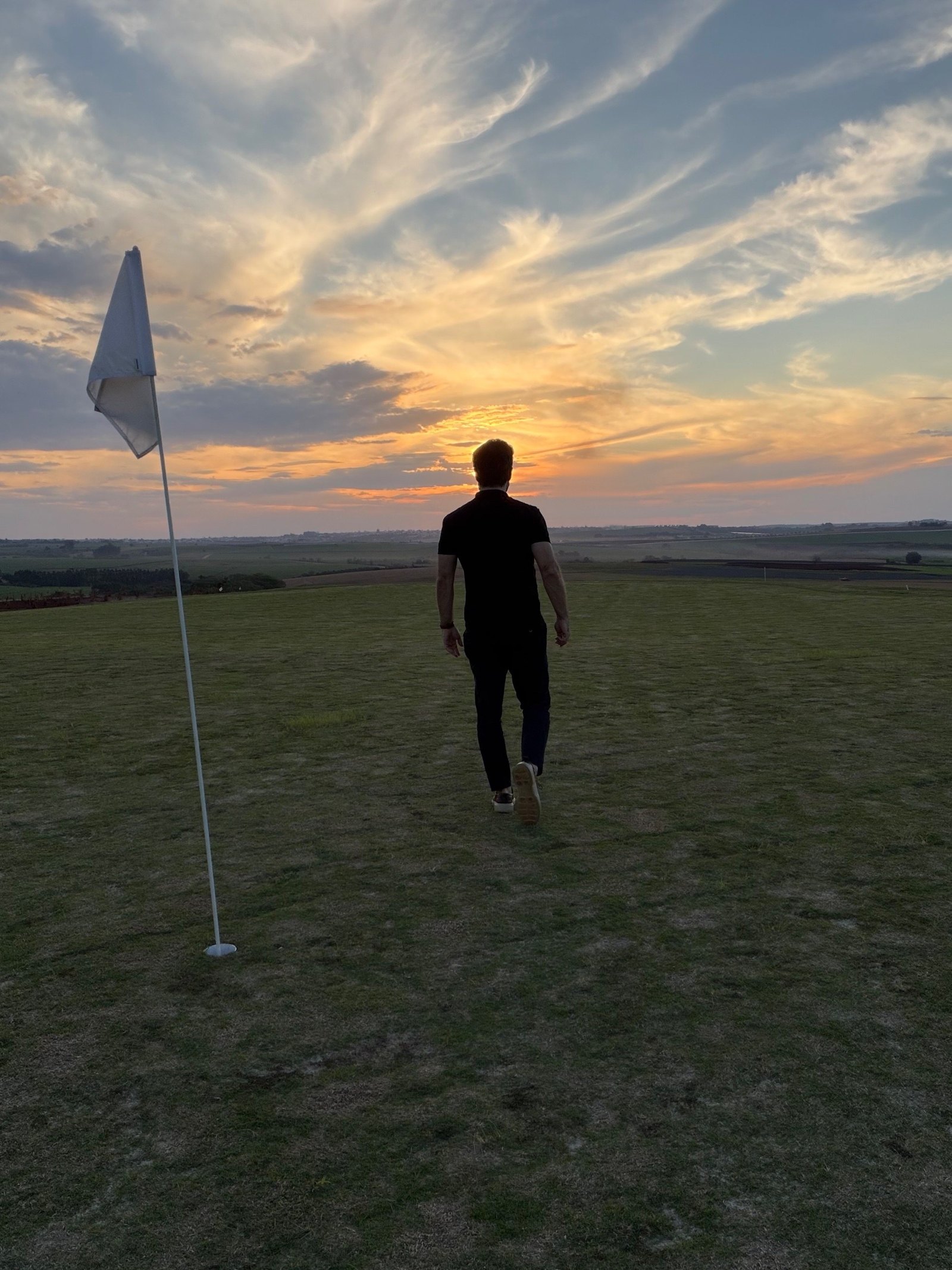 Tiago Bellini caminhando no campo do Heritage ao pôr do sol, com a bandeira do green em primeiro plano e a paisagem de Porto Feliz no horizonte